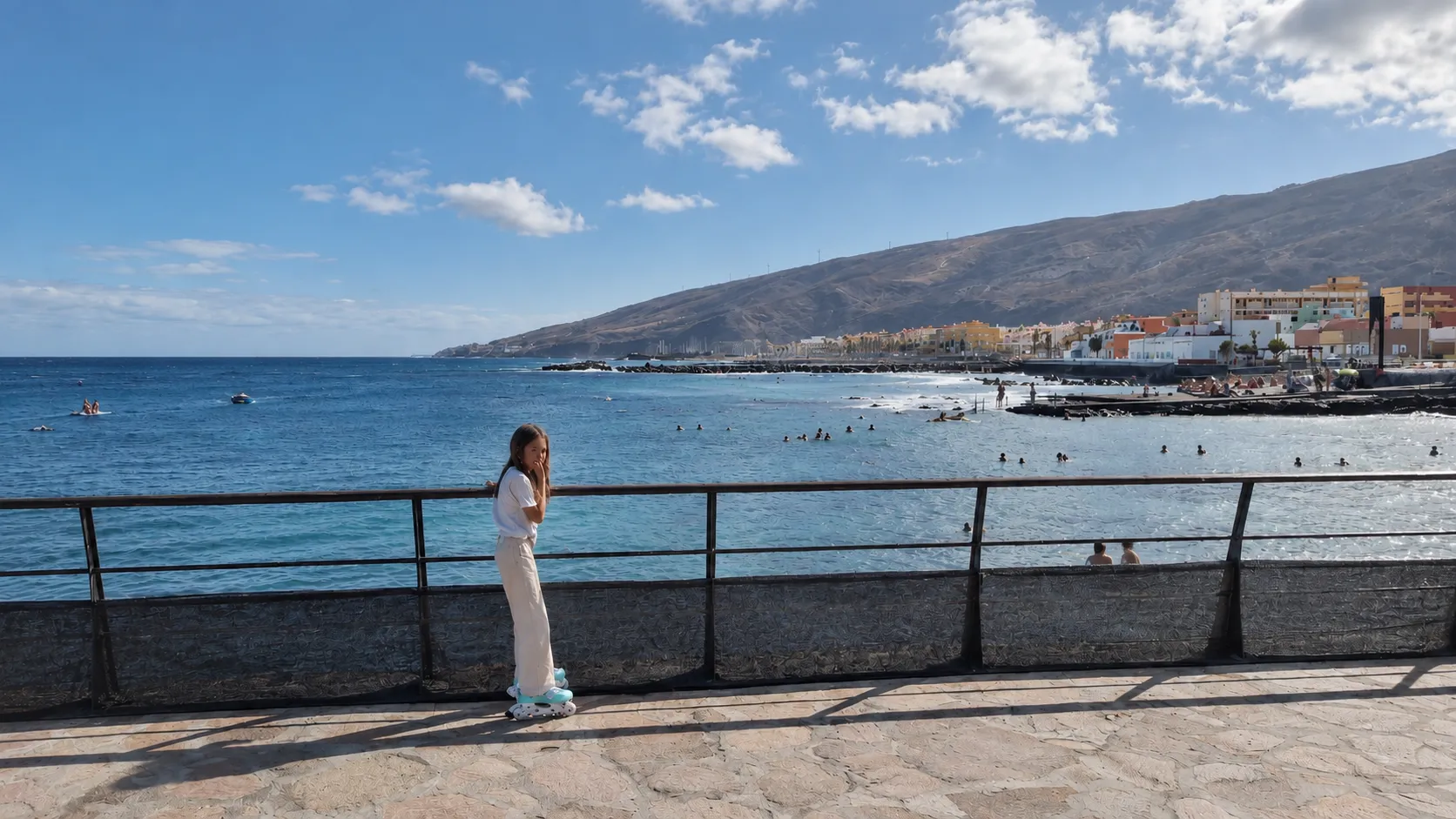 Girl on the seaside promenade, with swimmers in the sea and the coastline behind