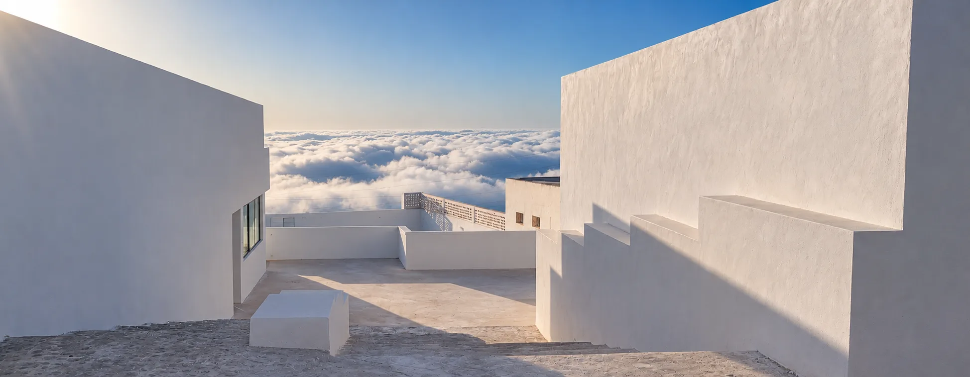 Weiße Mauern und Terrasse über einem Wolkenmeer, darüber blauer Himmel
